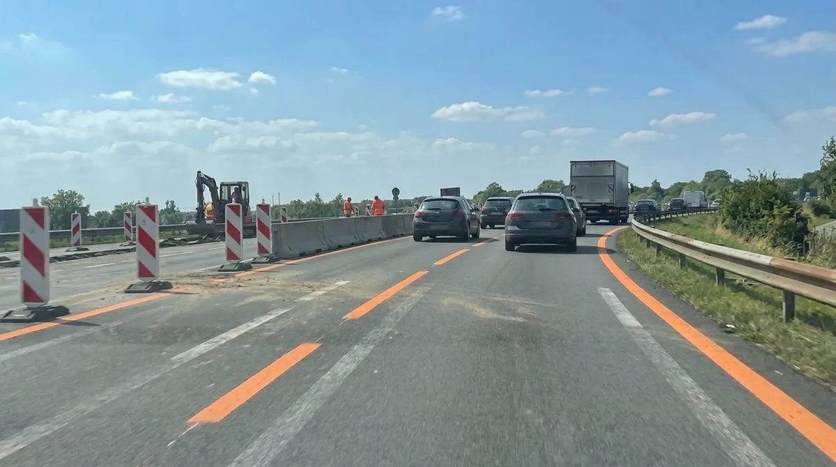 Vanuit de auto: op een autosnelweg met wegenwerken leiden oranje strepen het verkeer naar rechts, over de verbleekte witte strepen heen, met de werf links achter rood-witte bakens.